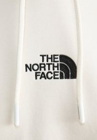 Felpa crema con cappuccio e due cordini bianchi, caratterizzata da un logo ricamato nero di "The North Face" e un design ad arco.