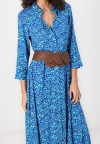 Blaues Blumenkleid mit Knopfleiste vorne, langen Ärmeln und einem braunen, oversized Gürtel mit kreisförmigen Ausschnitten und Nieten zur Verzierung.