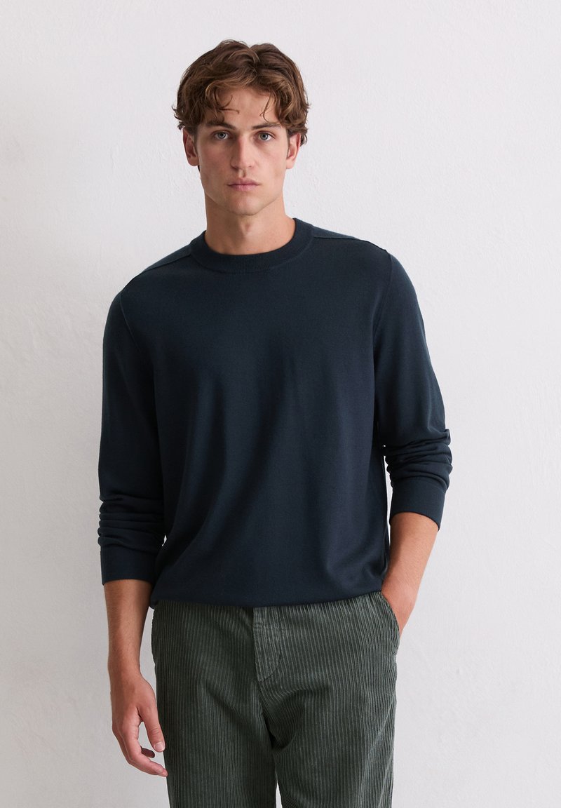 Pull en tricot bleu marine avec un col rond, des manches longues et des poignets côtelés, associé à un pantalon en velours côtelé vert olive avec des rayures verticales.