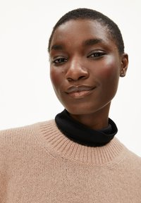 Lichtroze gebreide trui met een geribde hals, gedragen over een zwarte coltrui. Gladde texturen en een zachte, gezellige uitstraling.