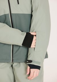 Softshelljacke in gedämpftem Grün mit einem kontrastierenden dunkelgrünen Streifen, ausgestattet mit einer Reißverschlusstasche und verstellbaren Bündchen. Glatte Textur, sportliche Passform.