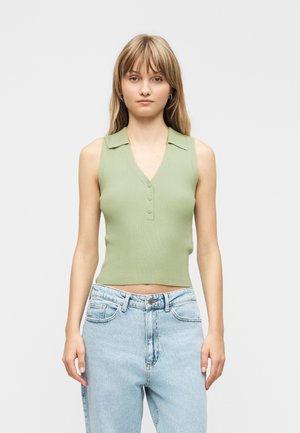 Jeune femme debout, portant un haut sans manches vert clair côtelé avec col et boutons, associé à un jean taille haute bleu clair.
