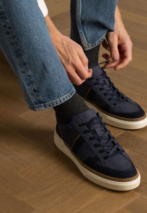 Marineblauwe sneakers van suède en textiel, met een witte zool en bruine accenten. Een persoon rijgt ze terwijl hij grijze sokken en een spijkerbroek draagt.