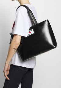 Glänzende schwarze Tragetasche mit stabilen Griffen. Verfügt über ein Etikett mit "Made with love" und "MOSCHINO"-Markierung. Glatte Textur, rechteckige Form.