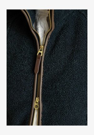 Mörk marinblå fleecejacka med brun dragkedja. Guldmetalldragkedjans dragtofs och tygfoder syns tydligt. Texturerad yta ökar värmen.