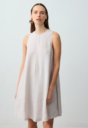 Frau mit langen braunen Haaren, die ein ärmelloses, hellgraues A-Linien-Kleid und blumige Creolen trägt, steht vor einem schlichten, hellen Hintergrund.