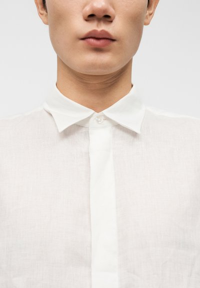 Gros plan sur le bas du visage et le cou d'une personne portant une chemise en lin blanche boutonnée avec un col pointu.