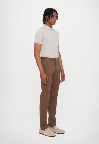 Polo a maniche corte grigio chiaro abbinato a pantaloni marroni su misura e scarpe slip-on beige. Design semplice, dettagli minimi, tessuto dalla consistenza liscia.