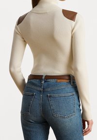 Osoba v krémovém pleteném roláku s hnědými ramenními záplatami a modrých denimových džínách s hnědým opaskem, zobrazená zezadu.