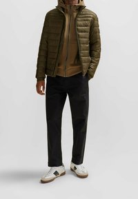 Olivově zelená péřová bunda s prošívaným vzorem, nošená přes hnědý zipový svetr. Kombinováno s černými kalhotami a bílými teniskami s hnědými akcenty.