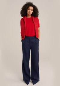 Punane kudumisvest ja ühtlane lühike vest, mis on sobitatud kõrge vöökohaga laia jalgadega tumesiniste pükstega. Kangas tundub pehme ja tekstuuriga viimistlusega.