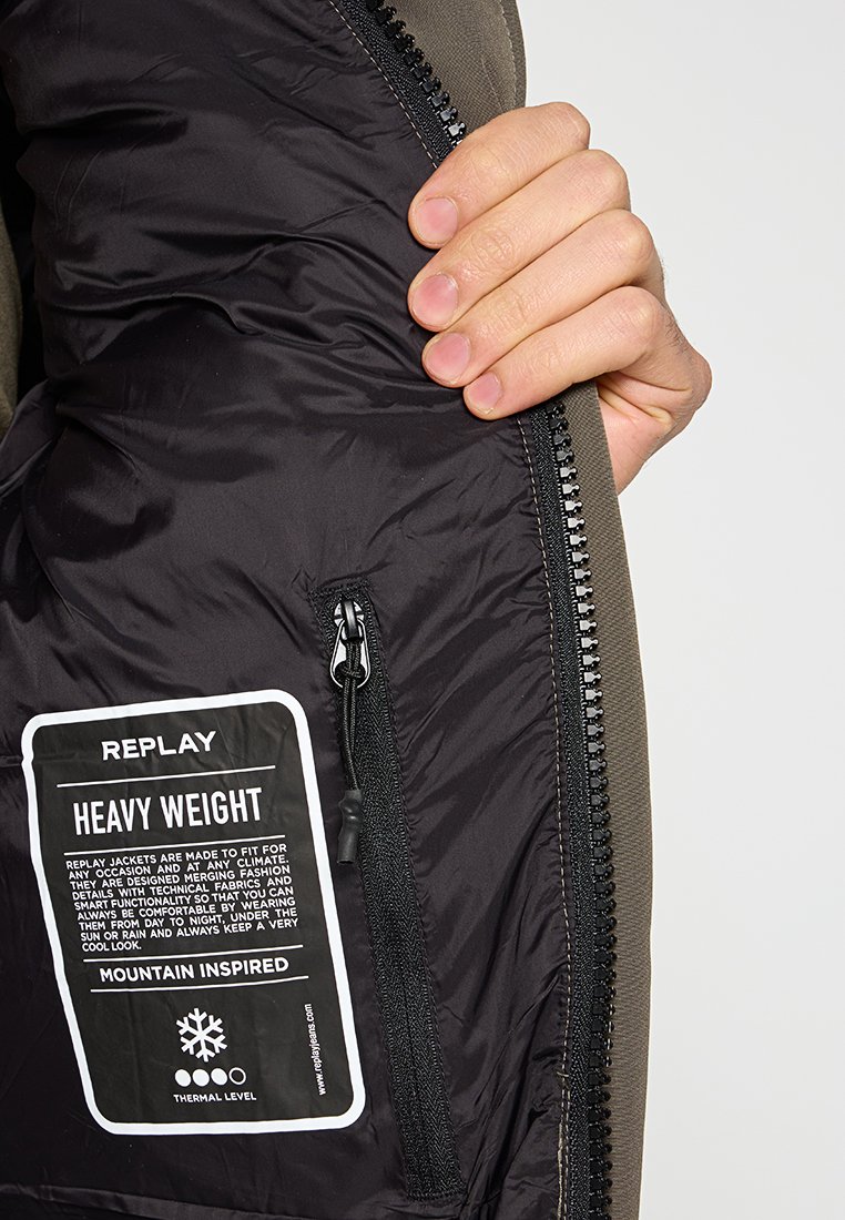 Die Innenseite der schweren Jacke verfügt über ein schwarzes Futter, eine Reißverschlusstasche und ein Etikett. Die Außenlage ist olivgrün und besteht aus strukturiertem Stoff.