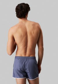 Costumi da bagno grigi con elastico in vita, bordi bianchi e un tessuto leggero e lucido; indossati da una persona vista da dietro.