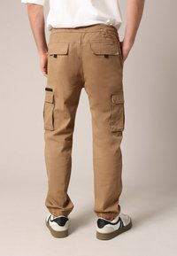 Pantalon cargo marron avec ceinture élastique, six poches et bas effilés, fabriqué en tissu léger. Présente des accents noirs sur les poches.