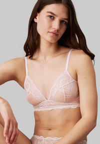 Bralette en dentelle de couleur rose doux avec des bonnets triangulaires, des bords festonnés délicats, des bretelles ajustables et une texture transparente, porté avec des strings en dentelle assortis.