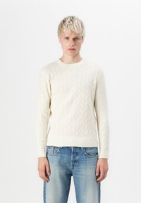 Cream cable knit sweater with a round neckline and ribbed cuffs, paired with light blue denim jeans. Simplistic and textured design.