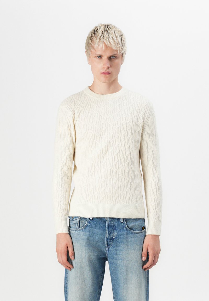 Cream cable knit sweater with a round neckline and ribbed cuffs, paired with light blue denim jeans. Simplistic and textured design.