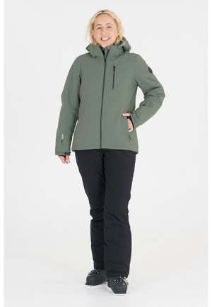 Grüne Softshelljacke mit einem Reißverschluss, verstellbarem Kapuze und zwei Vorderaschen. Getragen mit schwarzen Hosen und Winterstiefeln, auf weißem Hintergrund.
