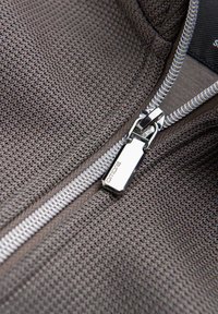 Gros plan sur un tissu gris texturé avec une fermeture éclair argentée brillante partiellement ouverte, montrant le curseur de la fermeture éclair gravé de "OMMRE".