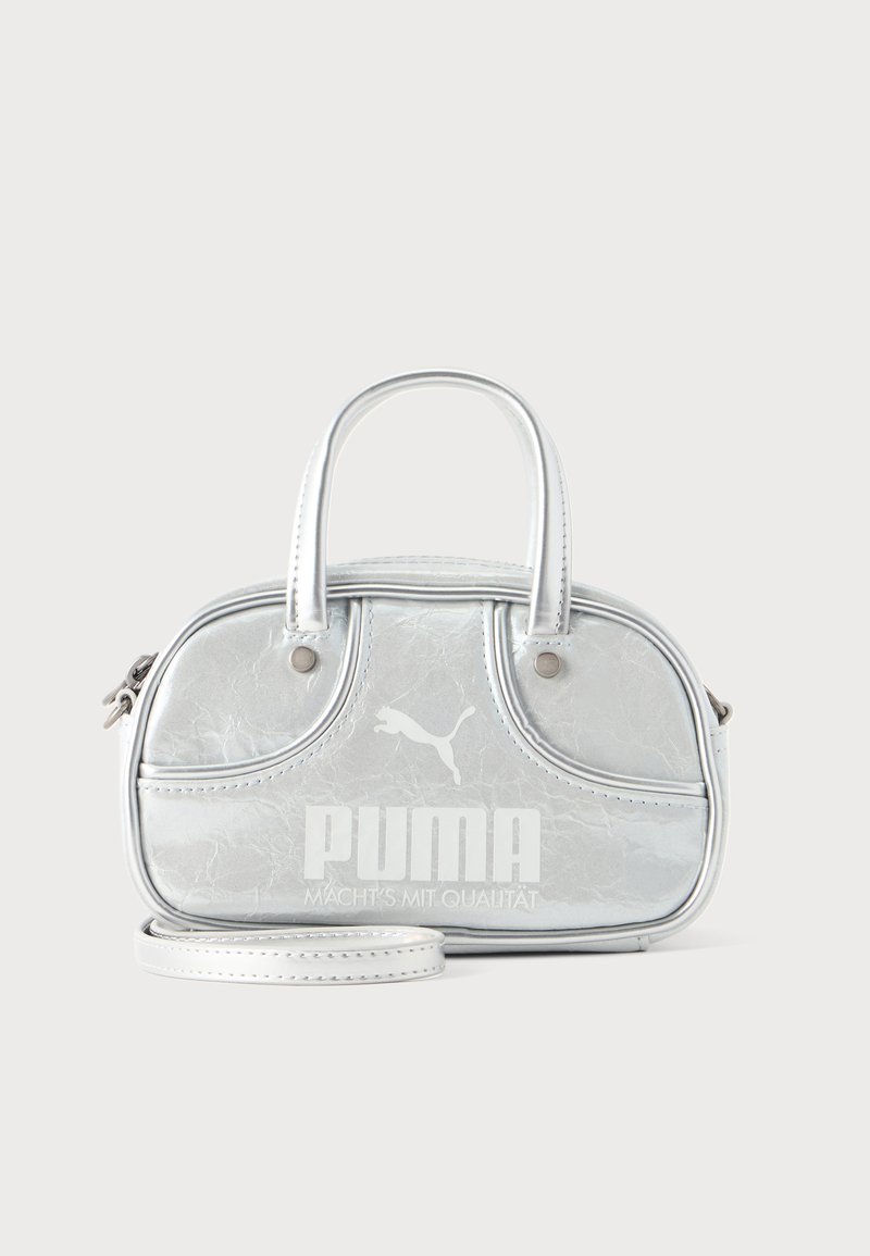 Μικρή μεταλλική ασημί τσάντα Puma με δύο χειρολαβές, μακρύ λουρί, λογότυπο μπροστά και κλείσιμο με φερμουάρ σε απλό ανοιχτόχρωμο φόντο.