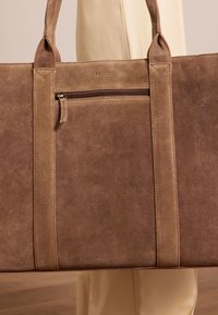 Bruine suède tote bag met gestructureerde vorm, bovenhandvatten, een buitenzak met rits en matte gouden rits hardware. Minimalistische branding, geperst aan de voorkant.
