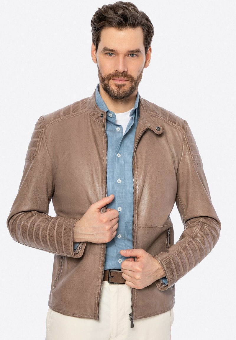 Giubbotto in pelle marrone con accenti a coste sulle spalle e chiusura con zip. Indossato sopra una camicia azzurro chiaro, caratterizzata da una texture liscia e una vestibilità su misura.