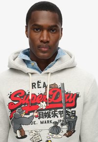 Felpa grigia con un grande grafico di cartone rosso che presenta un personaggio e un testo. I laccetti e il colletto in denim sono visibili sotto.