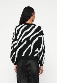 Femeie cu păr lung și creț, purtând un pulover negru cu dungi albe asemănătoare zebrei și pantaloni negri cu dungi subțiri, văzută din spate.