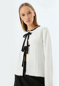 Witte cardigan met zwarte accenten, voorzien van een kabelbreisel ontwerp, een ronde hals en een gestrikte sluiting. Gelaagd textuur met een verkorte zoom.
