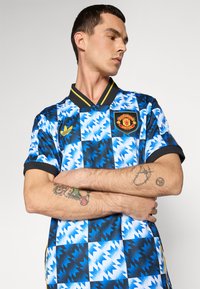 Blau- und schwarz gemustertes Sporttrikot mit Kragen, versehen mit einem gestickten Manchester United-Logo und gelben Akzenten.