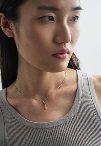 Colar dourado com pendente em forma de gota pendurado no pescoço de uma modelo; regata cinzenta texturada visível. Corrente delicada e detalhes de design minimalistas.