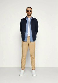 Casaco azul-marinho com botões, camisa azul clara, calças caqui e ténis brancos. O modelo usa óculos de sol pretos. Design simples, ajuste casual.