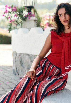 Kobieta siedząca na zewnątrz na białej kamiennej ławce, ubrana w czerwoną, bezrękawnikową bluzkę, spodnie w paski czerwono-granatowo-białe, z różowymi kwiatami w tle.