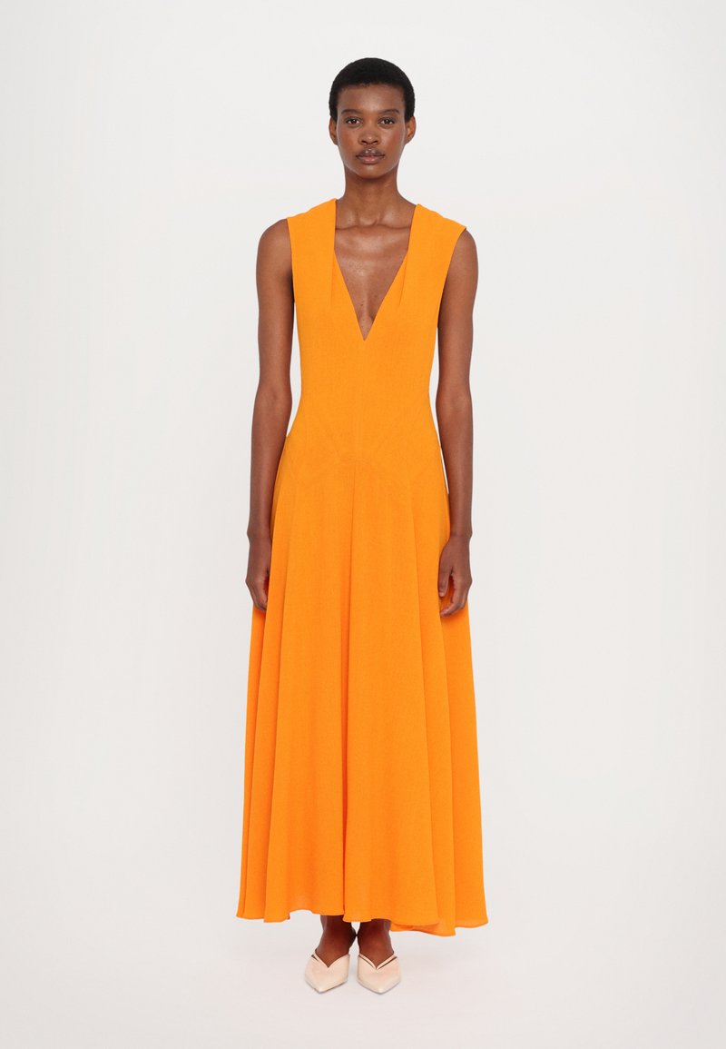 Long, sleeveless dress in bright orange fabric, featuring a deep V-neck and a flowing skirt, paired with light-colored shoes.