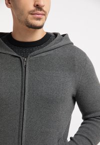 Grijze zip-up hoodie met een gestructureerde zwarte binnenkraag, voorzien van een capuchon en gemaakt van gladmateriaal met een aansluitend ontwerp.