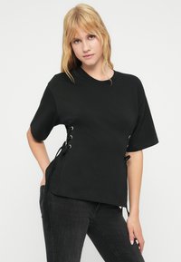 Μαύρο βαμβακερό μπλουζάκι με κοντά μανίκια, στρογγυλή λαιμόκοψη και ρυθμιζόμενα κορδόνια στο πλάι με ασημένιες μεταλλικές λεπτομέρειες.