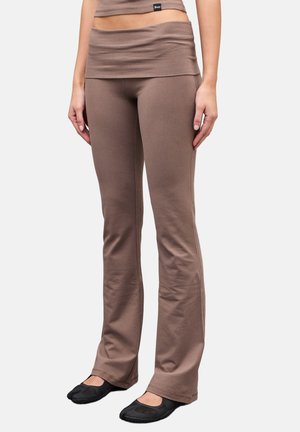 Mujer con pantalones marrones acampanados de talle alto y cinturilla ancha, y zapatos negros sin cordones, de pie contra un fondo liso.