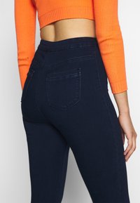 Jeans elásticos de color azul marino con una cintura alta, que cuentan con bolsillos traseros y una textura suave de denim oscuro. Suéter corto de canalé naranja visible.