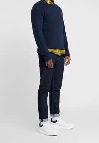 Marineblaues Langarmshirt über einem gelben Karohemd, kombiniert mit dunkelblauen Jeans und weißen Sneakers mit marineblauen Akzenten.