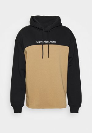 Tweedelige hoodie met zwarte bovenkant en mouwen, beige lijf, trekkoorden aan de capuchon en tekst 'Calvin Klein Jeans' op de borst.