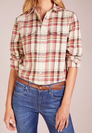 Femme portant une chemise à carreaux rouge, beige et verte avec manches retroussées, jean bleu et ceinture marron avec boucle dorée.