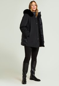 Sort dunjakke med falsk pelskrave, trykknapper og quiltet tekstur. Båret over sorte leggings og knæhøje sorte læderstøvler.