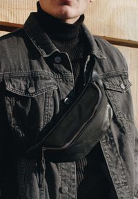 Black leather waist bag with a curved design and silver zipper, worn over a dark turtleneck and gray denim jacket, featuring silver hardware buttons.