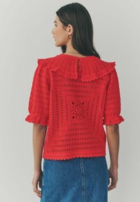 Haut au crochet rouge avec un col festonné, des manches courtes bouffantes et un détail dans le dos en forme de trou de clé. Le design texturé présente des motifs géométriques.