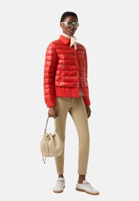 Rote Steppjacke mit kurzem Schnitt und horizontalen Nähten, kombiniert mit beigen Hosen und weißen Sneakern. Accessoires sind eine helle Handtasche und eine Sonnenbrille.