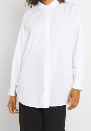 Personne portant une chemise blanche ample à manches longues boutonnée avec un col pointu et un pantalon noir sur un fond uni.