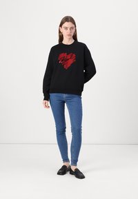 Svart sweatshirt med en röd hjärtdesign, med mjukt tyg, långa ärmar och ribbade muddiga ärmar, i kombination med blå åtsittande jeans och svarta loafers.