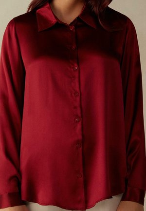 Mujer con blusa de satén rojo oscuro, mangas largas abotonadas, con cuello clásico y puños abotonados.