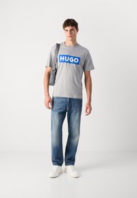 Pilkas trumpas rankovėmis marškinėliai su dideliu mėlynu "HUGO" logotipu, derinamas su šviesiai mėlynomis džinsomis ir baltomis sportbačiais. Modelis laiko pilką petinę rankinę.