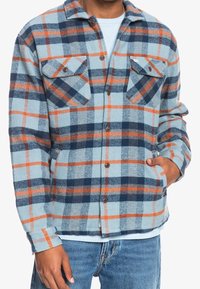 Man wearing light blue, navy, and orange plaid shirt jacket, with hand in side pocket, paired with blue jeans and a light blue undershirt.